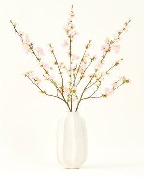 Silke kunstige blomsterbuketten Cherry blossom lys pink 94 cm har sarte pink kunstige kirsebær blomster i en hvid keramik vase, som tilføjer elegance mod en almindelig lys baggrund.