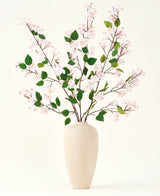 En beige keramikvase indeholder flere lange grene med Silk kunstige blomsterbuket af Peach Blossom pink og grønne blade, mod en almindelig hvid baggrund - perfekt til et kunstigt blomsterinteriør.
