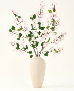En beige keramikvase indeholder flere lange grene med Silk kunstige blomsterbuket af Peach Blossom pink og grønne blade, mod en almindelig hvid baggrund - perfekt til et kunstigt blomsterinteriør.
