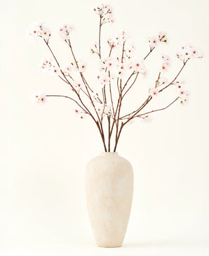 Silke kunstige blomsterbuketten Cherry blossom blush 114 cm har høje, sarte grene i en enkel beige vase, som tilføjer et blødt, elegant touch til dit interiør mod en almindelig, lys baggrund.