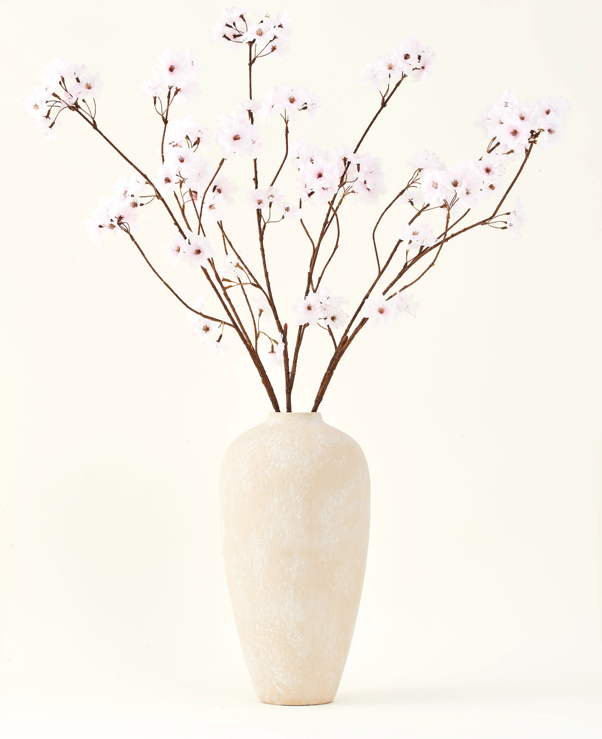 Silke kunstige blomsterbuketten Cherry blossom lys pink 114 cm står i en høj beige keramik vase med lange, tynde grene og sarte lys pink blomster, hvilket skaber en fredfyldt kunstig blomsteraccent mod en almindelig baggrund.