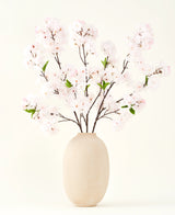 Silke kunstige blomsterbuketten Cherry blossom blush 119 cm viser sarte, lyserøde kirsebærblomster med grønne blade i en beige vase, ideel som en kunstig blomsterdekoration på en lys baggrund.
