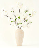 Silke kunstige blomsterbuketten Kirsebærblomstcreme 119 cm har elegante brune grene med bløde hvide falske blomster og grønne blade, vist i en enkel beige vase mod en almindelig lys baggrund.