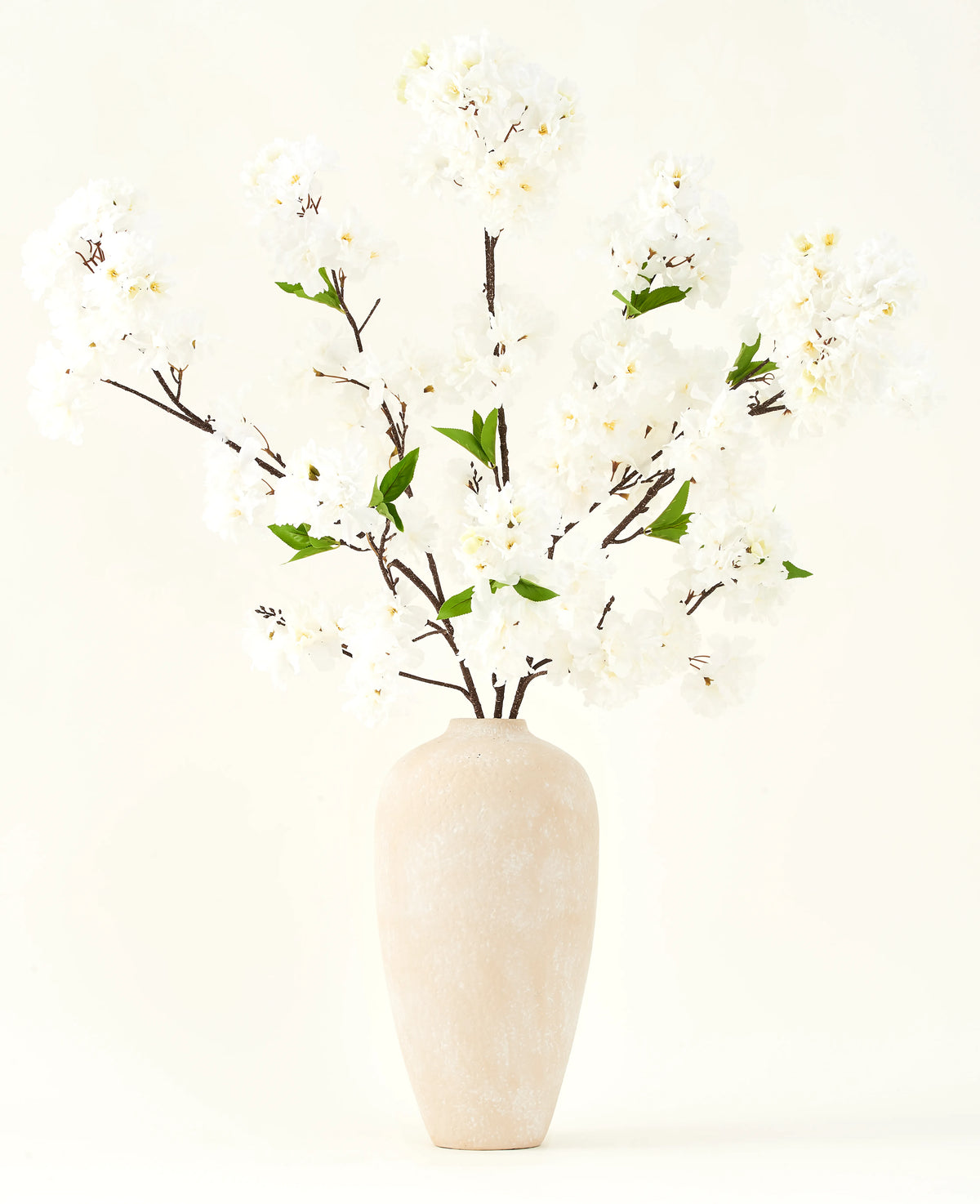 Silke kunstige blomsterbuketten Kirsebærblomstcreme 119 cm har elegante brune grene med bløde hvide falske blomster og grønne blade, vist i en enkel beige vase mod en almindelig lys baggrund.
