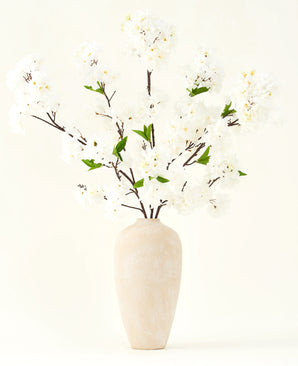 Silke kunstige blomsterbuketten Kirsebærblomstcreme 119 cm har elegante brune grene med bløde hvide falske blomster og grønne blade, vist i en enkel beige vase mod en almindelig lys baggrund.