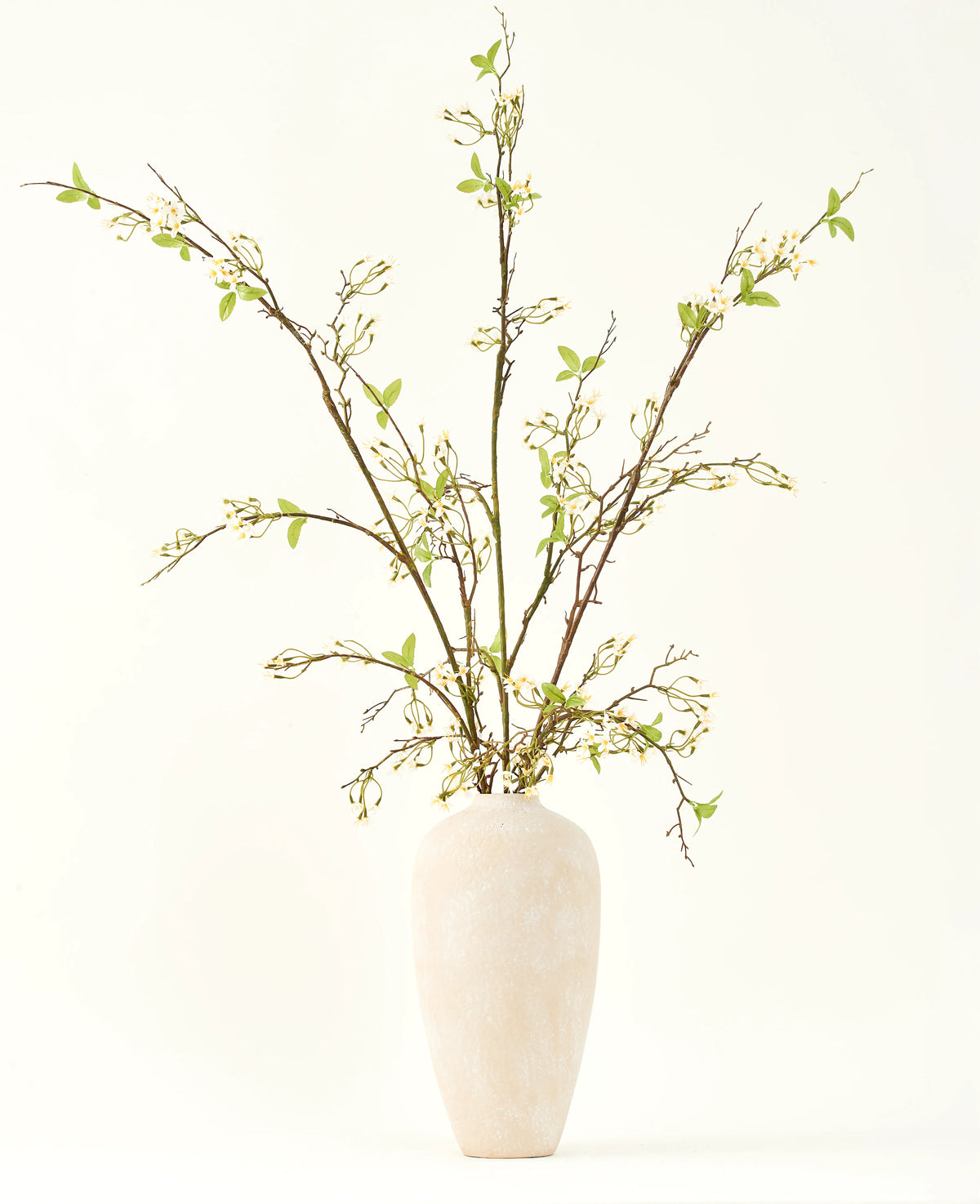 En høj, cremefarvet keramikvase indeholder Silke kunstige blomsterbuket Verbena creme med lange grene, små grønne blade og sarte hvide blomster, smukt arrangeret mod en almindelig lys baggrund.