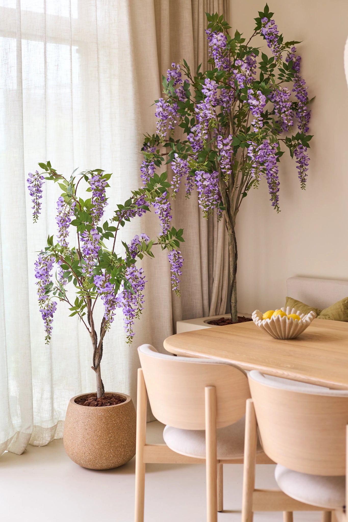 To kunstige Wisteria-planter på 150 cm, med deres overhængende lilla blomster, står ved siden af et spisebord i træ med lette stole. Blødt naturligt lys skinner gennem de gennemskinnelige gardiner bag planterne.