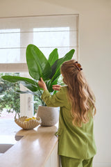 En kvinde med langt bølget hår, klædt i et grønt jakkesæt, plejer en 90 cm Strelitzia Real Touch Deluxe kunstig plante på en disk ved siden af en skål med citroner i et lyst rum med et vindue.