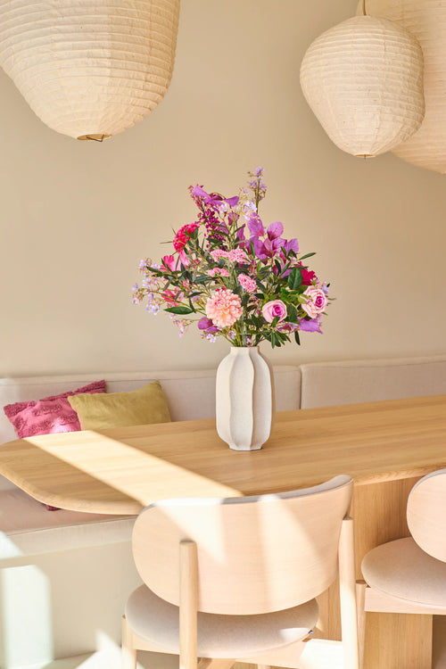På et lyst spisebord i træ står en hvid vase med Purple Dawn kunstige blomsterbuket i silke, sprængfyldt med levende lyserøde og lilla nuancer. Bagved fuldender en polstret bænk med puder og to hængende papirlanterner scenen, der gennemsyrer rummet med den rolige atmosfære inspireret af Purple Dawn.