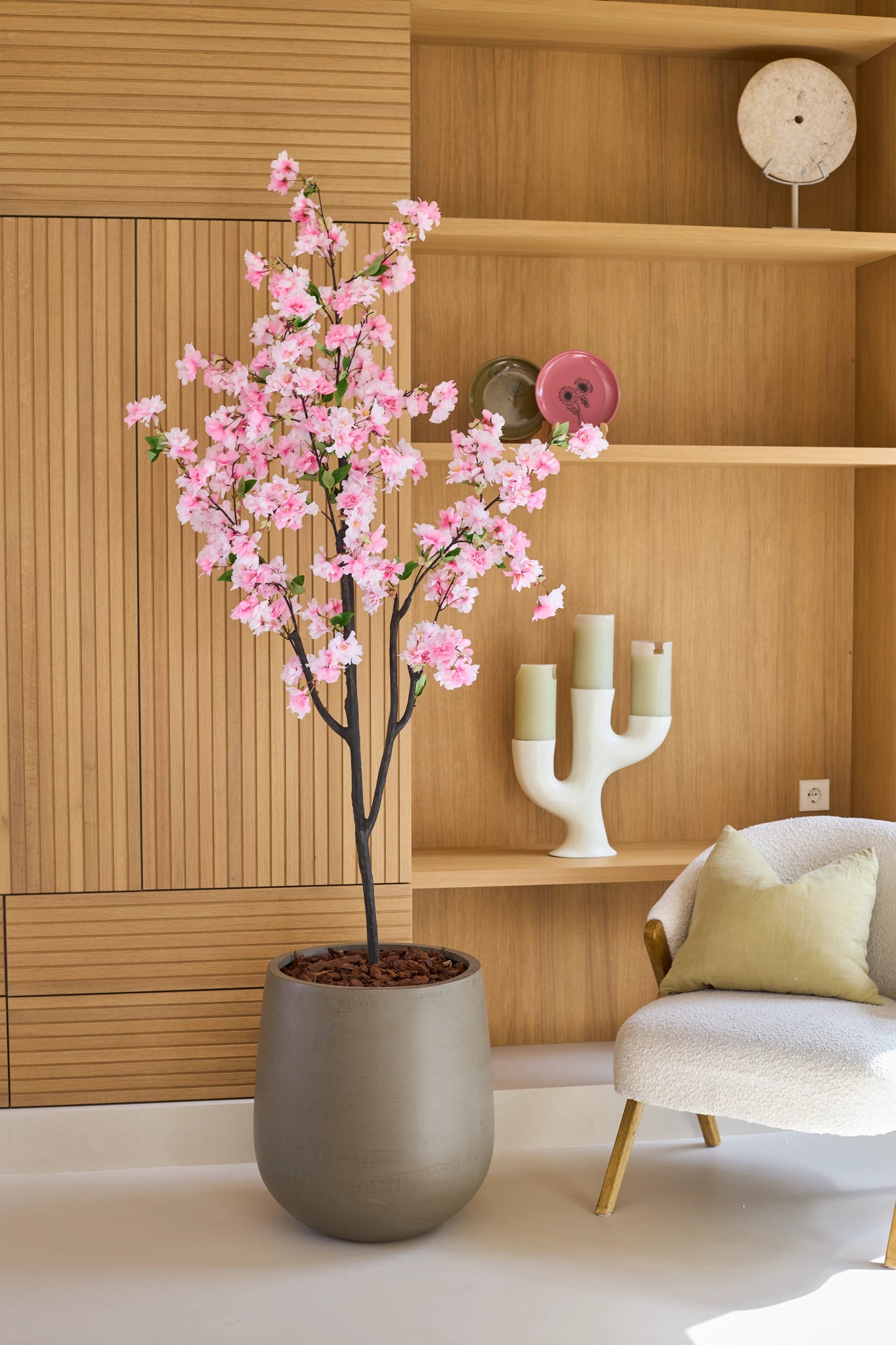 En kunstig kirsebærblomst 180 cm pink, et pottetræ med lyserøde blomster, står ved siden af en hvid stol med hynder i et moderne rum med træhylder og dekorative genstande.