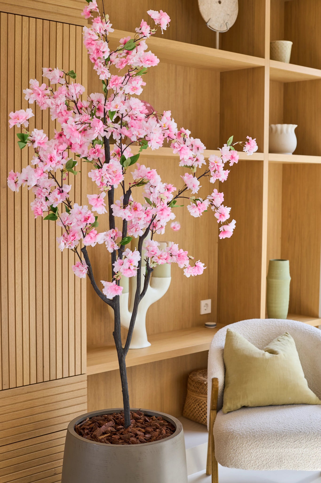The Artificial Cherry Blossom 180 cm pink, med sine sarte lyserøde blomster, står i en moderne stue med træhylder, keramik, en beige lænestol med en grøn pude og en minimalistisk indretning.