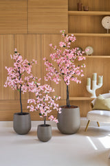 Tre kunstige bougainvilleaer på 180 cm, lyserøde og med lyserøde blomster, står foran væghylder i træ, ved siden af en moderne hvid stol med hynde.