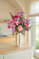 På en køkkenø af lyst træ står der en hvid vase med 'Purple Dawn' buket af kunstige silkeblomster - bestående af lyserøde og lilla kunstige blomster - med vindue og beige køkkenskabe i baggrunden.