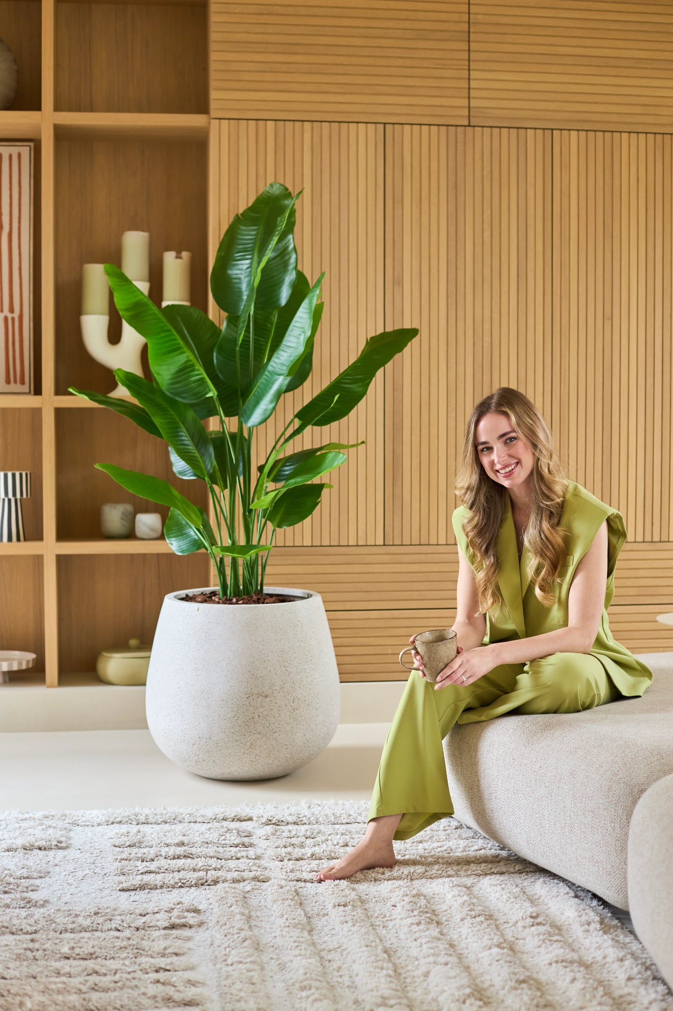 En kvinde i et lysegrønt outfit sidder barfodet på en sofa, smilende og holder et krus. Ved siden af hende står kunstplanten Strelitzia Real Touch Deluxe 150 cm, mens træplanker og dekorative genstande giver baggrunden et varmt udtryk.