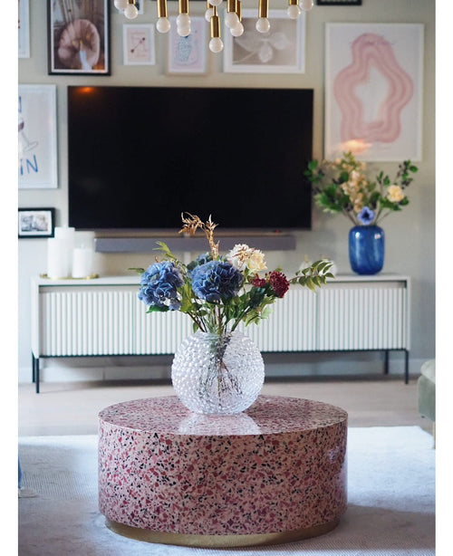 En moderne stue har et terrazzo sofabord med en glasvase med blomster, et tv på væggen, indrammet kunst og fotos, en hvid skænk og Artificial Hydrangea Deluxe 57 cm lilla som et elegant blåt blomsterarrangement i baggrunden.