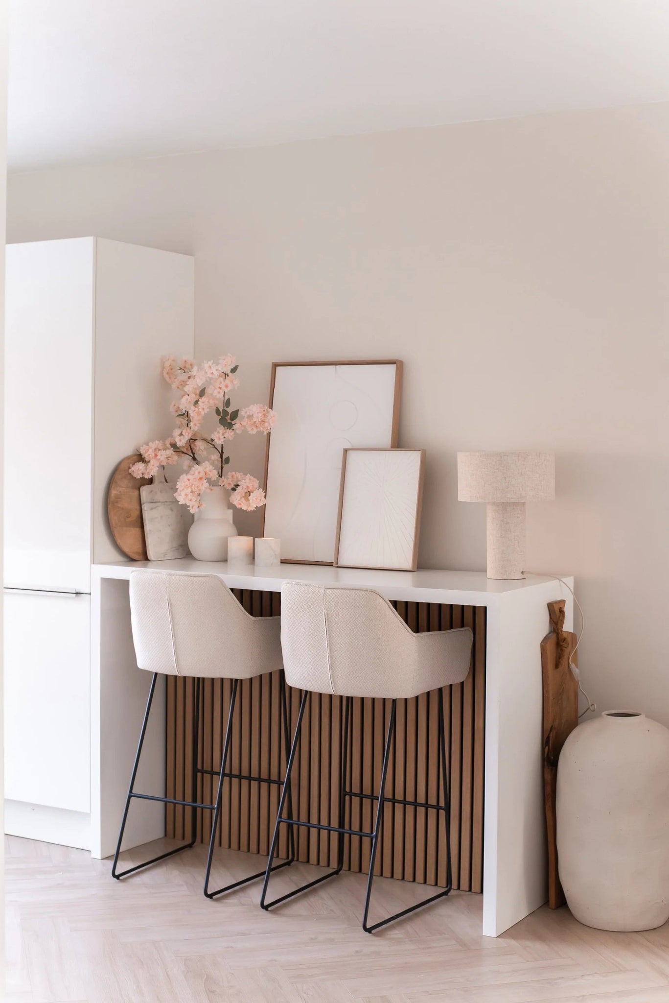 Een minimalistische thuisbar met twee beige krukken met kussens, een witte toonbank, ingelijste kunst, een tafellamp, neutraal getinte decoratie en Kunst Kersenbloesem 100 cm licht roze in vazen tegen een lichtgekleurde muur.