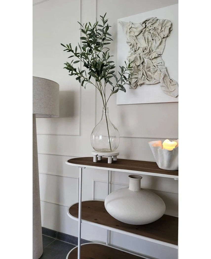 Et minimalistisk konsolbord viser en klar vase med den kunstige olivengren 109 cm, en rund hvid keramikvase og en hvid skål med LED-lys. Struktureret hvid vægkunst hænger over den, og en gulvlampe sidder i nærheden.
