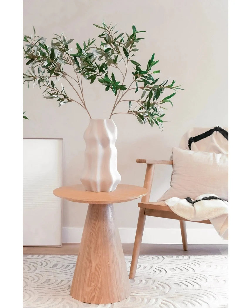 En moderne stue med en træstol med en let hynde, et rundt sidebord og en hvid skulpturel vase med kunstigt olivenblad 90 cm UV, hvilket skaber et minimalt rum med bløde neutrale toner og en subtil middelhavsatmosfære.