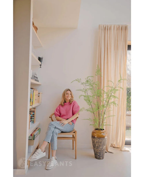 En kvinde i lyserød sweater og jeans sidder på en træstol ved siden af en 120 cm kunstig bregneplante, med reoler og et vindue med beige gardiner i et lyst, moderne rum.