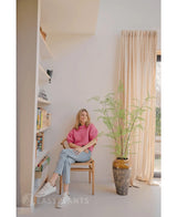 En kvinde i lyserød sweater og jeans sidder på en træstol ved siden af en 120 cm kunstig bregneplante, med reoler og et vindue med beige gardiner i et lyst, moderne rum.
