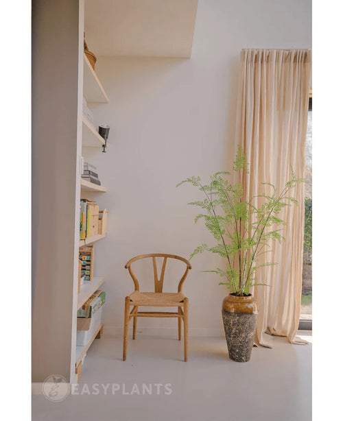 Ved siden af en 120 cm høj kunstbregne i en krukke står en træstol, nær cremefarvede gardiner og en væg med hylder fyldt med bøger og dekorationer. Sollys oplyser det minimalistiske rum, hvor stilfulde stueplanter som Art Fern tilføjer et strejf af naturinspireret ro.