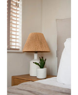 Et natbord med en hvid lampe med en vævet beige skærm og den kunstige sukkulentplante 20 cm står ved siden af en seng med hvidt og beige sengetøj, nær et vindue med træpersienner.