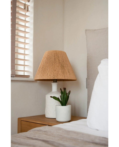 Et natbord med en hvid lampe med en vævet beige skærm og den kunstige sukkulentplante 20 cm står ved siden af en seng med hvidt og beige sengetøj, nær et vindue med træpersienner.