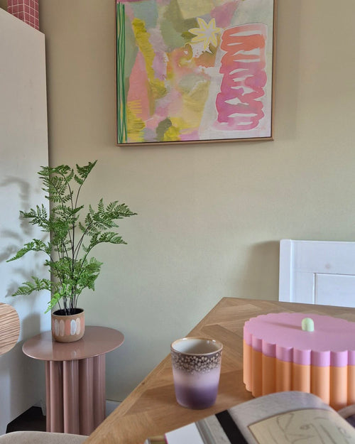 I et rum med pasteltema er der en 85 cm kunstig bregneplante på et lyserødt sidebord, en lilla og guld kop, en lyserød skål med en scalloped kant, en åben notesbog og på væggen et abstrakt maleri med grønne, pink og gule toner.