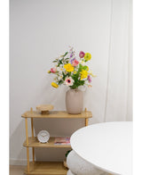 En lys træhylde viser en beige vase med Artificial Ranunculus Real Touch Yellow 60 cm blomster, et hvidt ur og en træbakke. En del af et rundt hvidt bord vises i forgrunden mod en almindelig hvid væg.