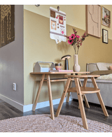 Et moderne stuehjørne med et lyst træ sofabord med den kunstige blomst Anemone grene 56 cm pink i en pink vase, sammen med en lampe og bøger. Ved siden af er der en beige sofa, fremhævet af vægkunst og en gallerilignende fototavle på den gule væg.