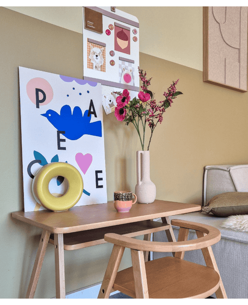Et træskrivebord med tilhørende stol viser et blåt fugleprint, kunstig blomst Anemone forgrenet 56 cm pink, en rund gul ornamentik og en lille kop. Ovenpå hænger en opslagstavle med fotos og sedler ved siden af en beige sofa.