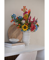 En kunstig nellikeblomst 70 cm blå i en hvid keramikvase står på et hvidt skrivebord ved siden af stablede bøger og en lille dekorativ skål, med en rund træbakke lænet op ad væggen bagved.