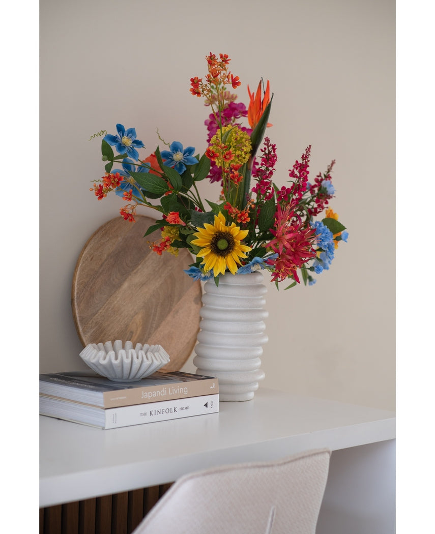 En kunstig nellikeblomst 70 cm blå i en hvid keramikvase står på et hvidt skrivebord ved siden af stablede bøger og en lille dekorativ skål, med en rund træbakke lænet op ad væggen bagved.