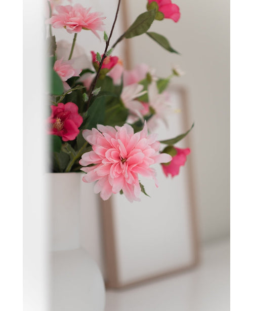 Et nærbillede af den kunstige blomst Dahlia 68 cm pink i en hvid vase, med en svag fotoramme i baggrunden på en hvid overflade.