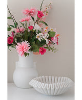 En Artificial Flower Dahlia 68 cm pink i en hvid vase står ved siden af en hvid keramik skål med flæser på en hvid overflade, mod en almindelig lys baggrund.