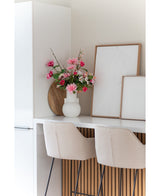 Et moderne hvidt køkken med to beige barstole, en Artificial Flower Dahlia 68 cm pink i en hvid vase på disken og to indrammede abstrakte kunstværker op ad væggen.