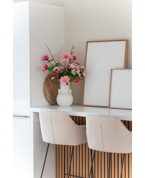 Et moderne hvidt køkken med to beige barstole, en Artificial Flower Dahlia 68 cm pink i en hvid vase på disken og to indrammede abstrakte kunstværker op ad væggen.