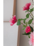 Et nærbillede af den kunstige blomst Rhododendron Azalea 73 cm fuchsia, med levende lyserøde kunstige blomster med grønne blade, mod en blød neutral baggrund og en del af en træramme synlig.