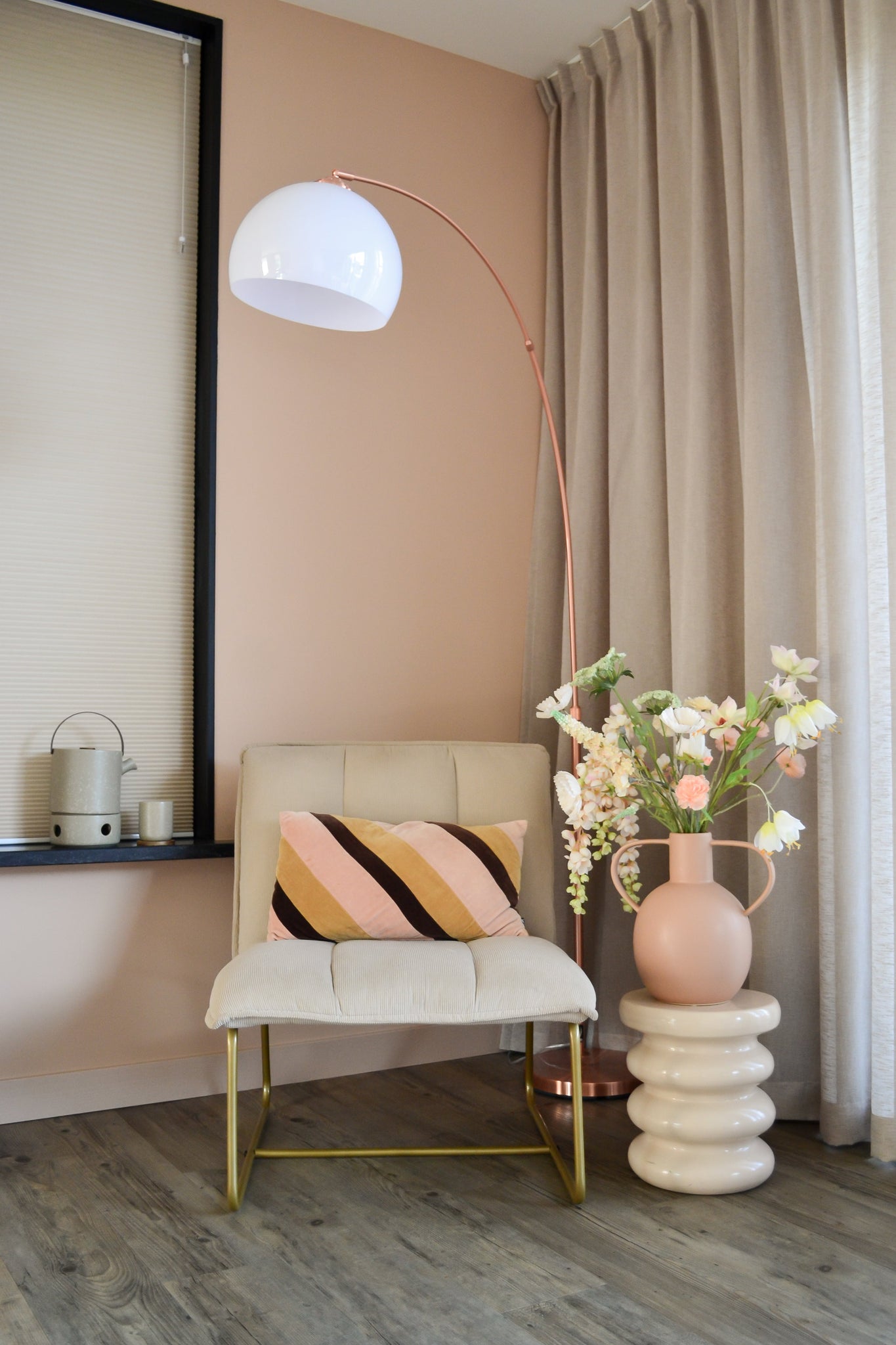 Et moderne hyggehjørne med en cremefarvet stol med en stribet hynde, en høj bueformet gulvlampe, beige gardiner, et trægulv og en rund vase med Artificial Flower Fritillary 94 cm hvide blomster på et skulptureret sidebord.