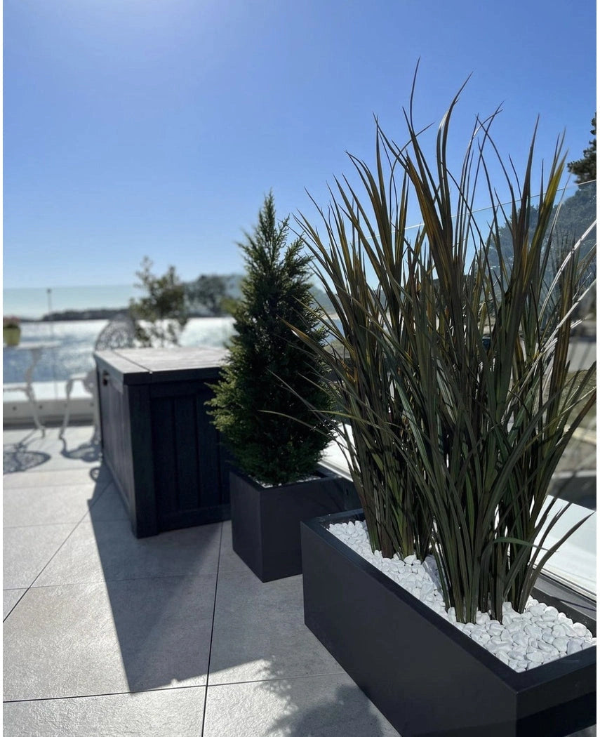Kunstgræs Bordeaux UV 120cm i rektangulære plantekasser med hvide sten dekorerer en moderne udendørs terrasse mod en baggrund af blå himmel og træer i det fjerne.