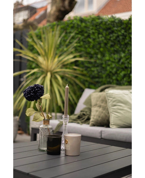 Et moderne udendørsbord med sort overflade viser en vase med en mørk blomst, en lysestage af glas og to kopper. Bagved står en sofa med grønne hynder overfor en frodig grøn væg lavet af Kunsthaag måttemix 50x50 cm.
