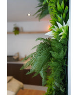 Et nærbillede af frodig grøn kunstig hæk Friskhed 100x100 cm UV kunstige planter pryder en lodret indervæg, med en sløret baggrund, der viser neutrale møbler og en lille hængende plante.