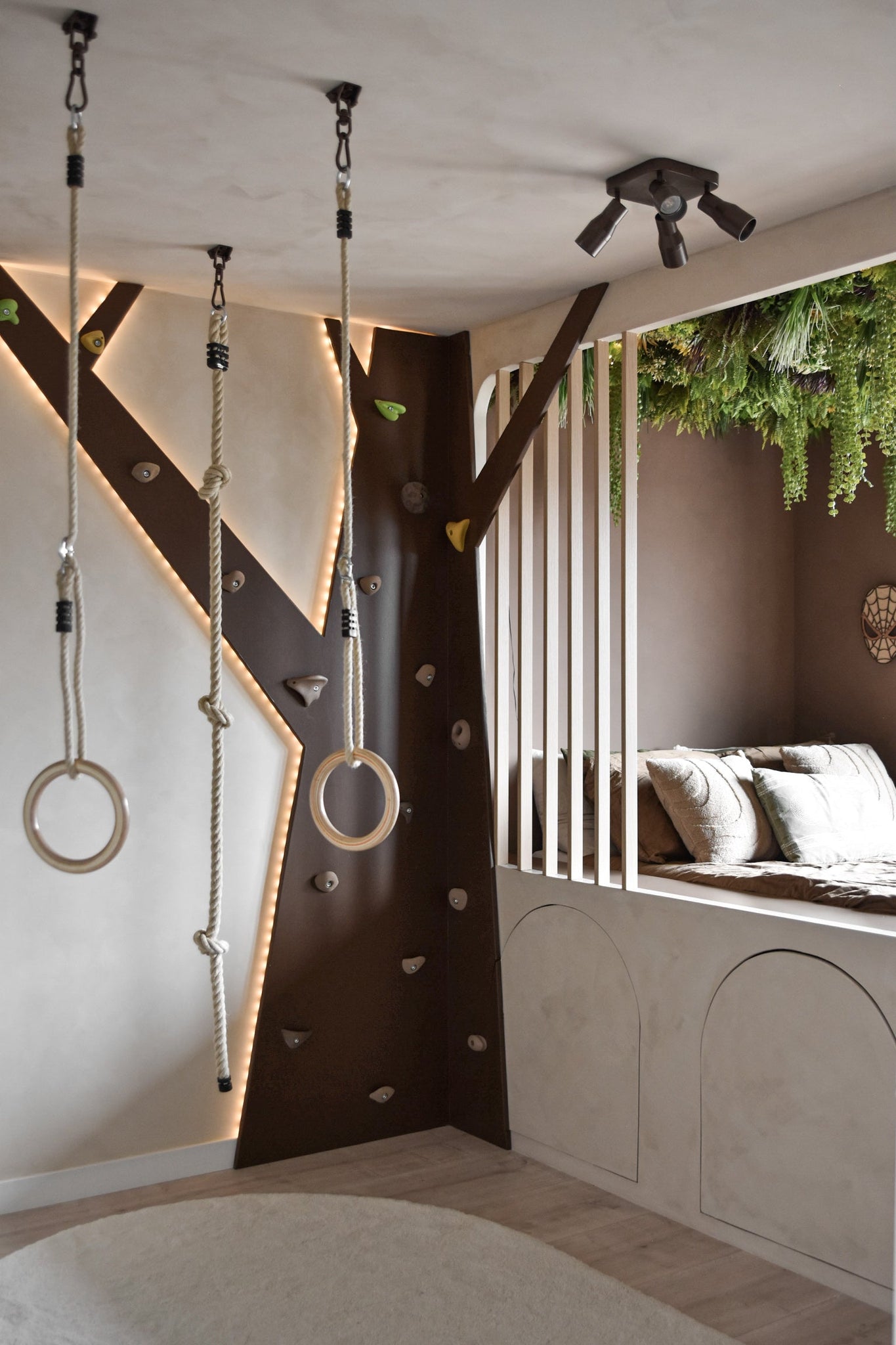 Een moderne kinderspeelkamer heeft een klimwand, gymringen, touw en een gezellige leeshoek met kussens en verticale latten. Kunsthaag Lush Deco Deluxe 100x100 cm UV voegt groen toe aan het plafond en vult de neutrale tinten en speelse verlichting van de kamer aan.