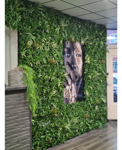 Et 100x100cm Kunsthaag Savanne brandhæmmende panel med frodigt grønt omgiver et stort sort/hvidt ansigtsportræt i et indvendigt rum med flisebelagte loftpaneler og en lille skranke til venstre.