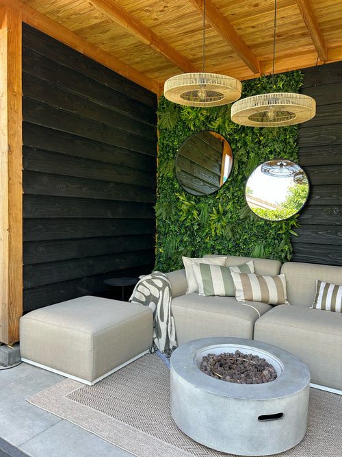 Moderne udendørs opholdsområde med en beige sofa, stribede hynder og et rundt bålbord. To runde spejle hænger på en kunstig hæk Volta 100x100 cm UV plantevæg under et træloft og flettede hængelamper.