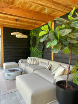Et hyggeligt udendørs opholdsområde med en beige sofa, stribede hynder, en rund bålplads, en stor potteplante og en træpergola. Væggen er dekoreret med grønt med Artificial Haag Volta 100x100 cm UV for ekstra stil.