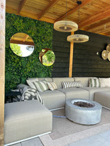 En hyggelig terrasse med en beige sofa, stribede hynder, bålplads og vævede tæpper. UV grøn fra Kunsthaag Volta 100x100 cm hænger på væggen, runde spejle og flettede lamper under en træpergola.