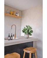 En moderne bordplade med en messinghane, to sæbedispensere, den kunstige tobaksplante Fiddle Leaf UV 45 cm potteplante og to træskamler. En flydende hylde med krukker og flasker er monteret på en lys væg.
