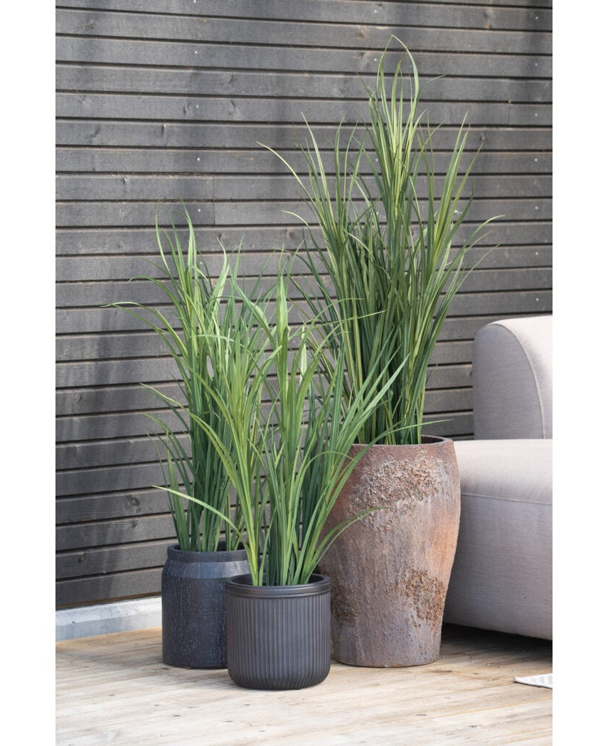 Tre kunstgræsgrønne UV 150 cm kunstige planter med lange, smalle blade står på et træterrasse ved siden af en lysegrå udendørs sofa, mod en mørk træspaltevæg.