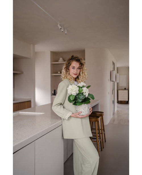 En kvinde med krøllet blond hår, klædt i et lysegrønt outfit, står i et moderne køkken med en Artificial Hydrangea White UV 35 cm plante i hænderne – med cremede hvide blomster – og smiler til kameraet.